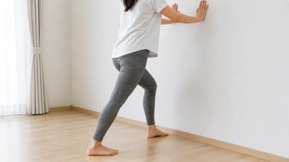 壁に両手をつき、片足を後ろに伸ばしてふくらはぎをストレッチしている日本人女性の後ろ姿(顔は写っていません)